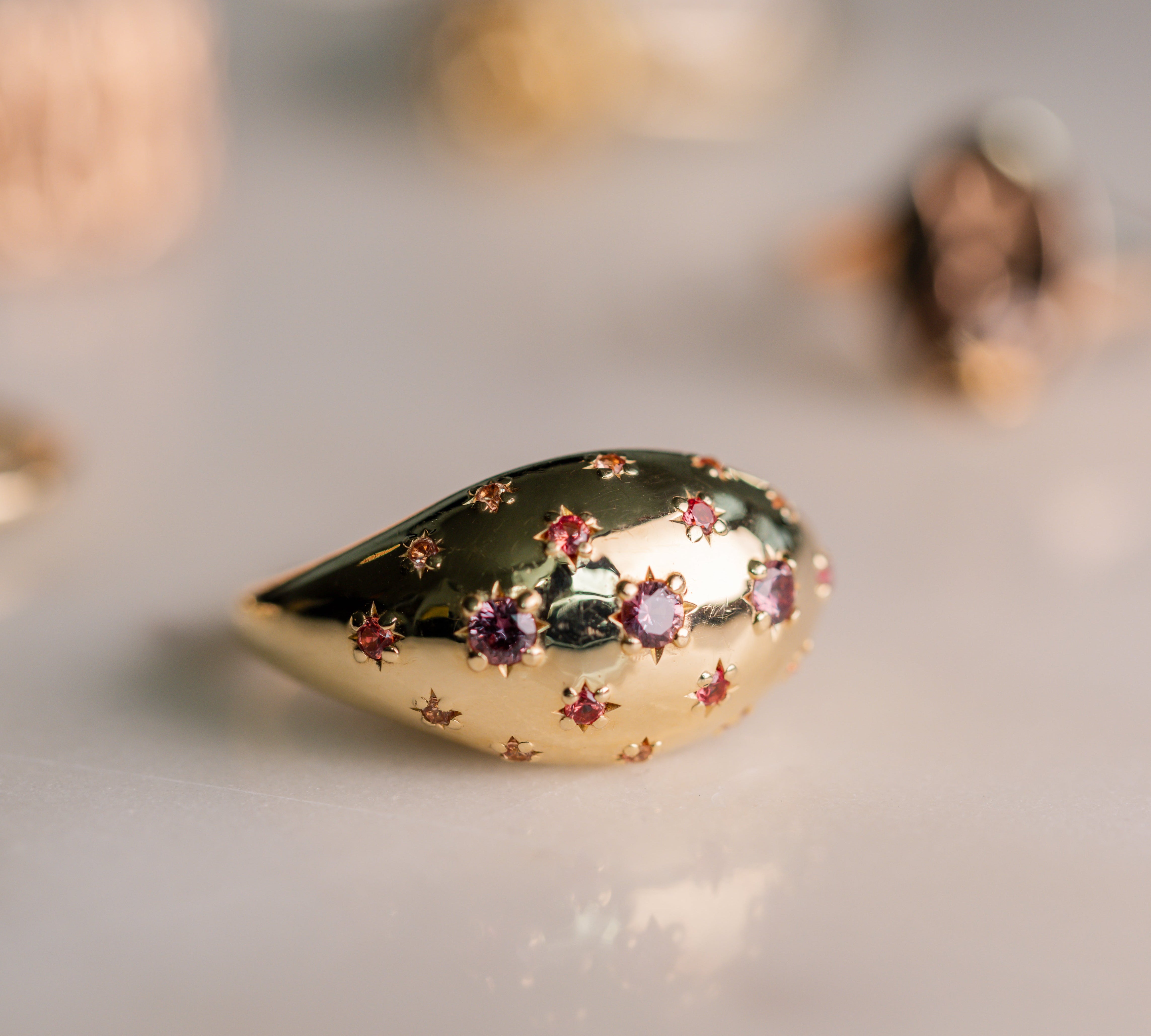 A gold teardrop-shaped ring with star-shaped engravings and small purple gemstones, photographed on a light gray surface with a soft-focus background.