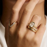 A close-up of a person's hand wearing multiple gold rings, including a ring with a cushion-cut clear gemstone and smaller clear stones, against a soft white and black background.