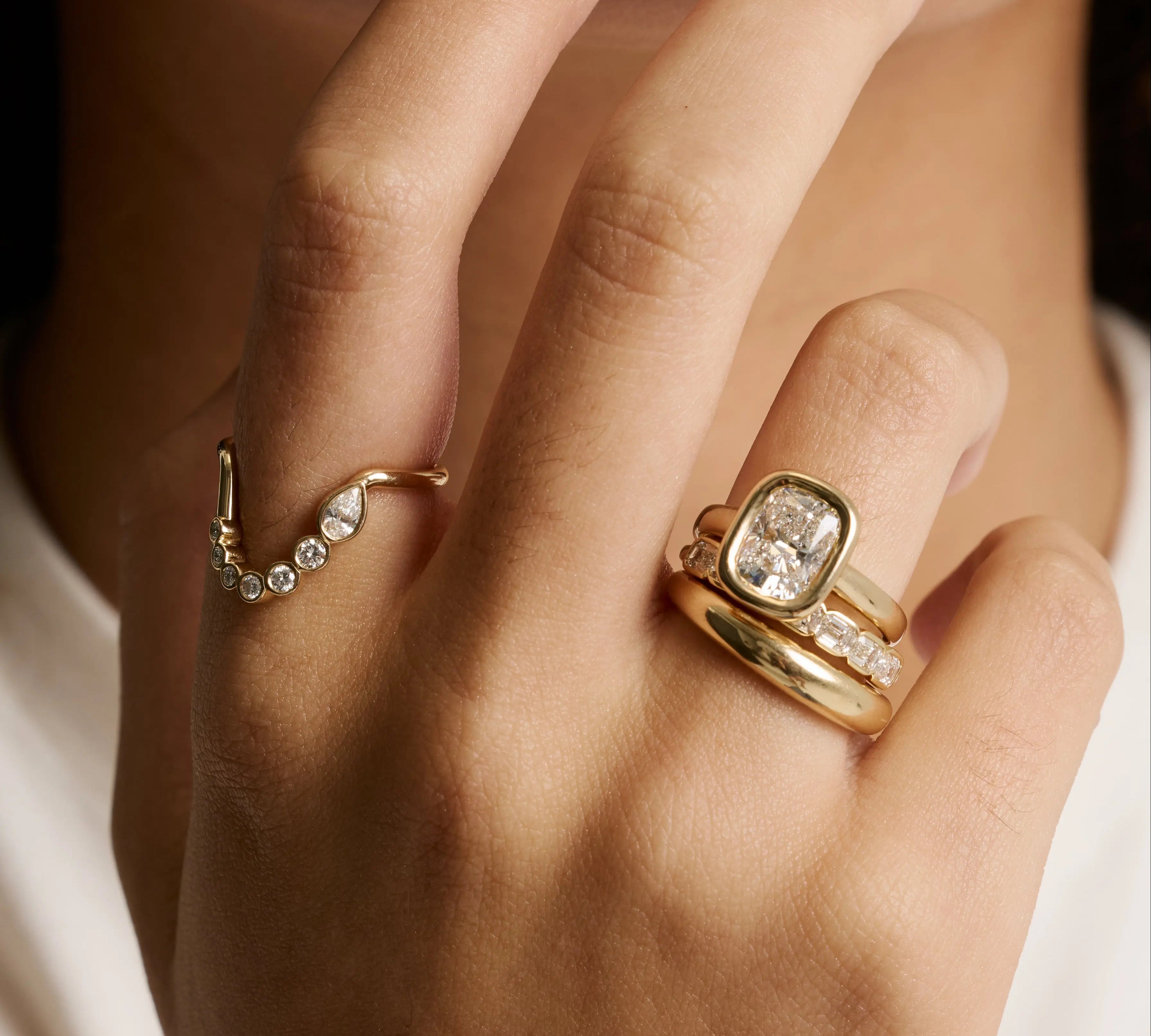 A close-up of a person's hand wearing multiple gold rings, including a ring with a cushion-cut clear gemstone and smaller clear stones, against a soft white and black background.