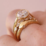 A close-up of a gold ring with a large round clear gemstone and smaller marquise-cut stones, worn on a finger against a soft pink background.