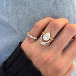 A close-up of a hand wearing a gold ring set featuring a pear-cut diamond center stone surrounded by baguette and round diamonds, alongside a thin diamond band, set against light blue denim.