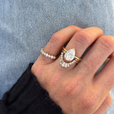A close-up of a hand wearing a gold ring set featuring a pear-cut diamond center stone surrounded by baguette and round diamonds, alongside a thin diamond band, set against light blue denim.
