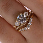 A close-up of a gold ring set on a finger, featuring a large oval-cut clear center stone with smaller round stones, alongside a curved diamond-accented band.