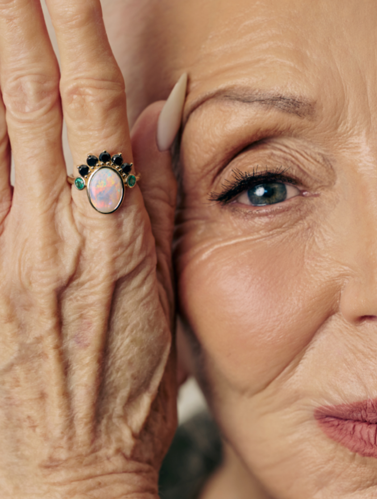 Close-up portrait of an older woman with one hand raised to her face, wearing a ring with a pink opal center and dark green and black stones on a gold band.