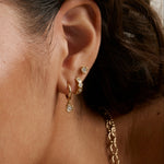 A close-up of a model's ear showcasing a stack of gold earrings, including a huggie hoop with a small clear stone dangle, a curved cluster of three small gold studs, and a larger gold post stud. A gold chain necklace is partially visible.
