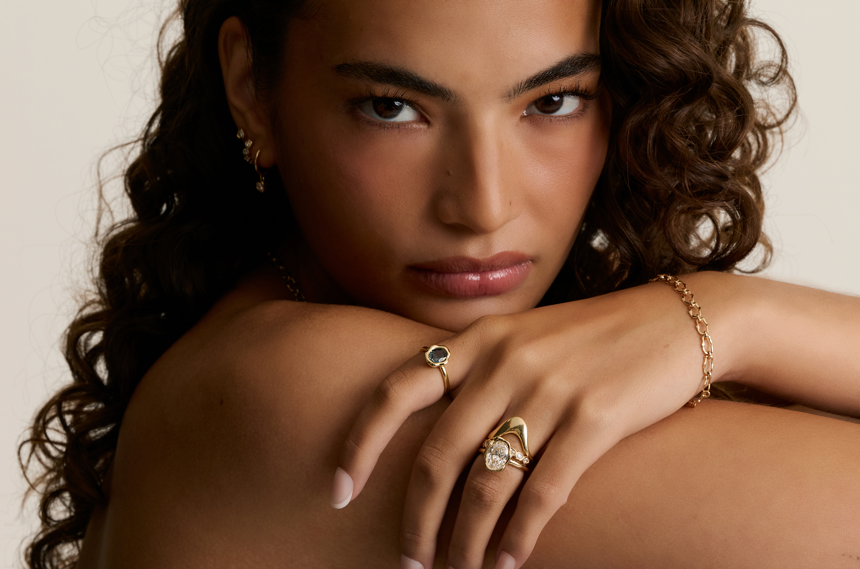 Close-up of a woman with curly brown hair wearing gold rings and a gold chain bracelet, resting her head on her hand against a light beige background.