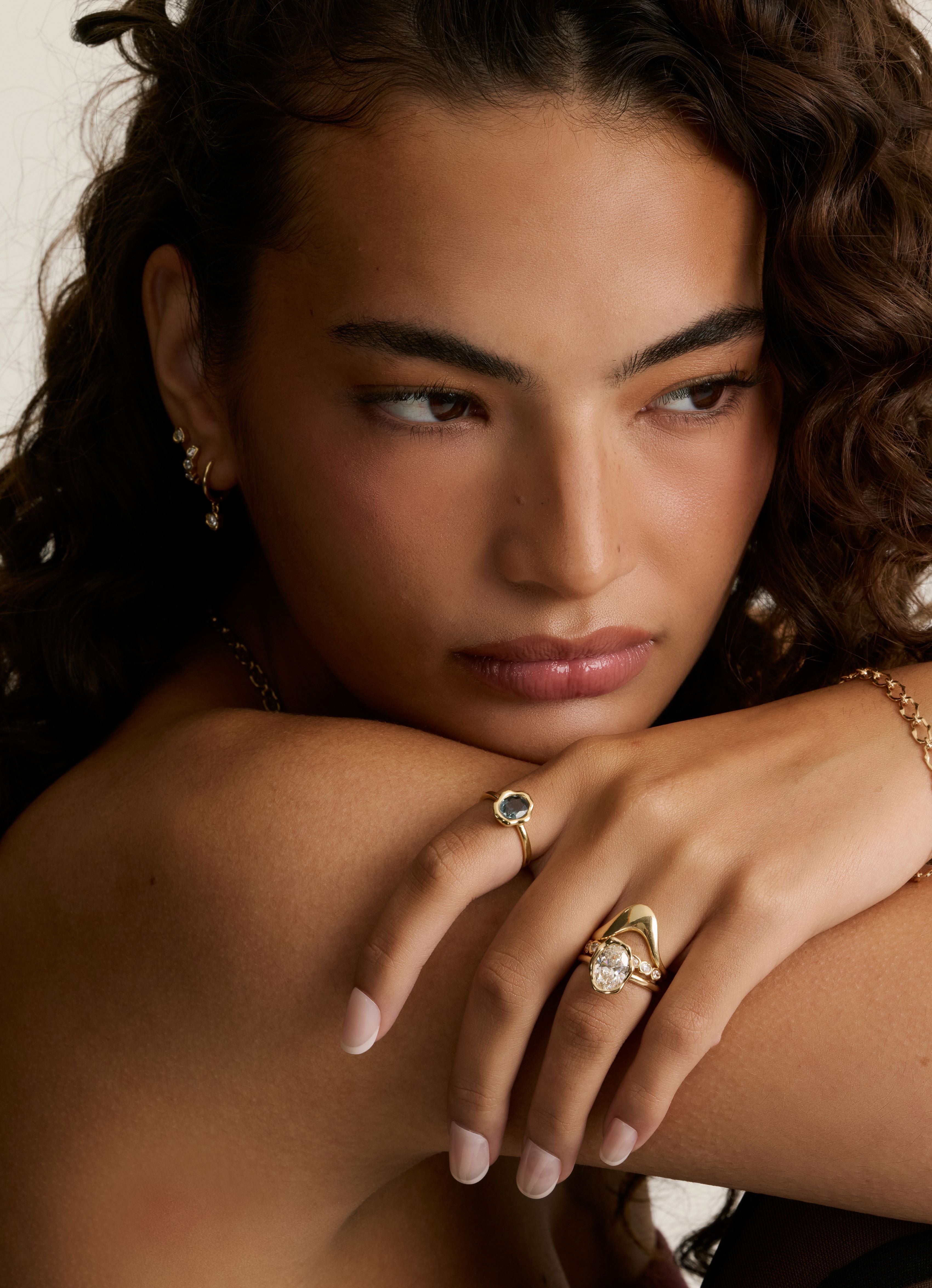 A close-up lifestyle shot of a woman with curly brown hair wearing gold rings and a gold chain bracelet. She is resting her chin on her hand, showcasing the jewelry against a warm, neutral background.