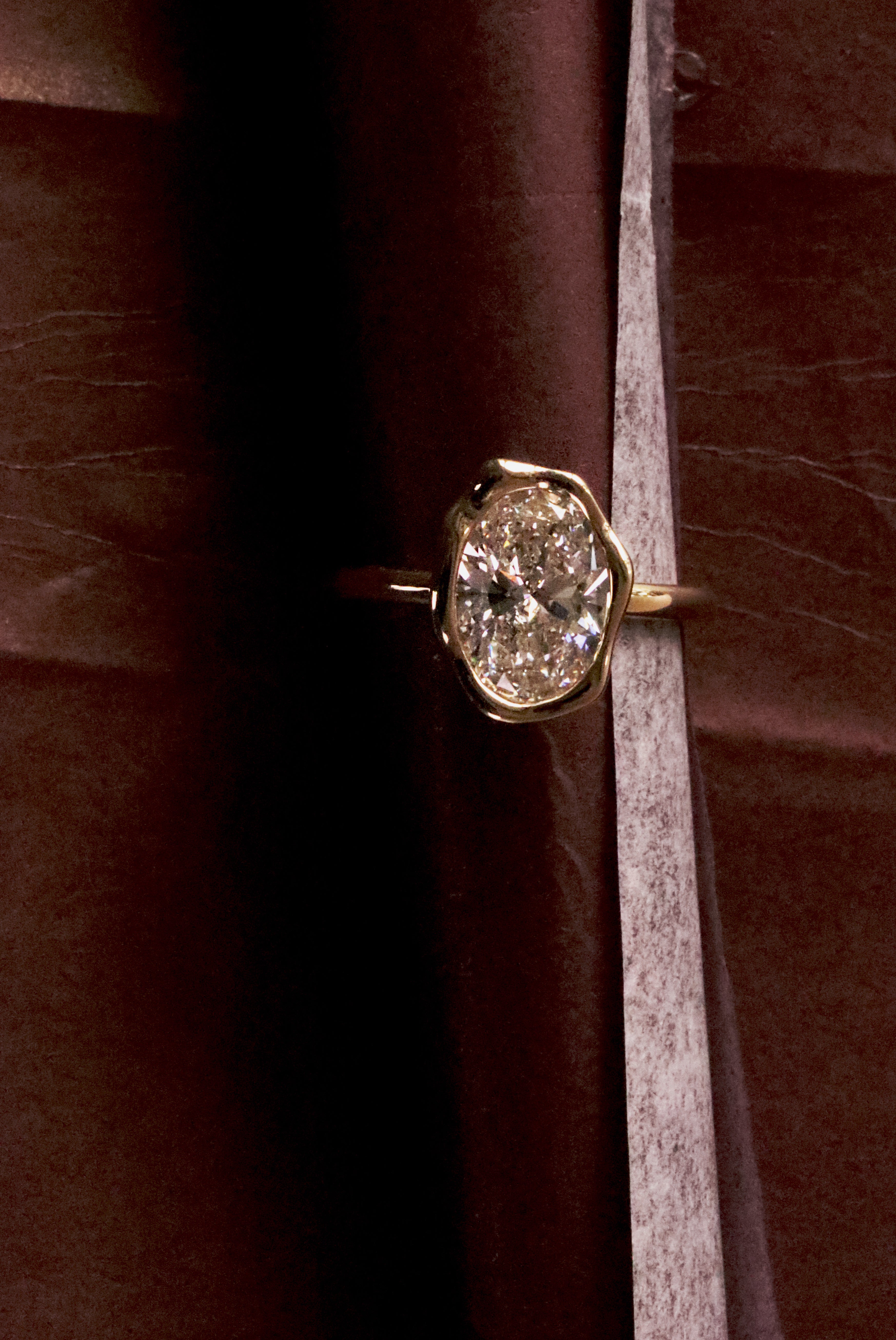 A yellow gold ring with a large round brilliant-cut diamond set in an abstract bezel setting, resting horizontally on a dark brown velvet fabric background.