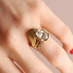 A close-up of a hand wearing a gold ring with a large oval white diamond set in an abstract bezel and small accent stones on a slim band, set against a soft beige background.