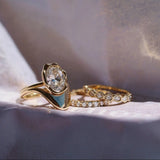 A gold ring set with a large oval clear gemstone and a light blue inlay, displayed alongside two gold bands with small clear stones on a soft lavender fabric background.