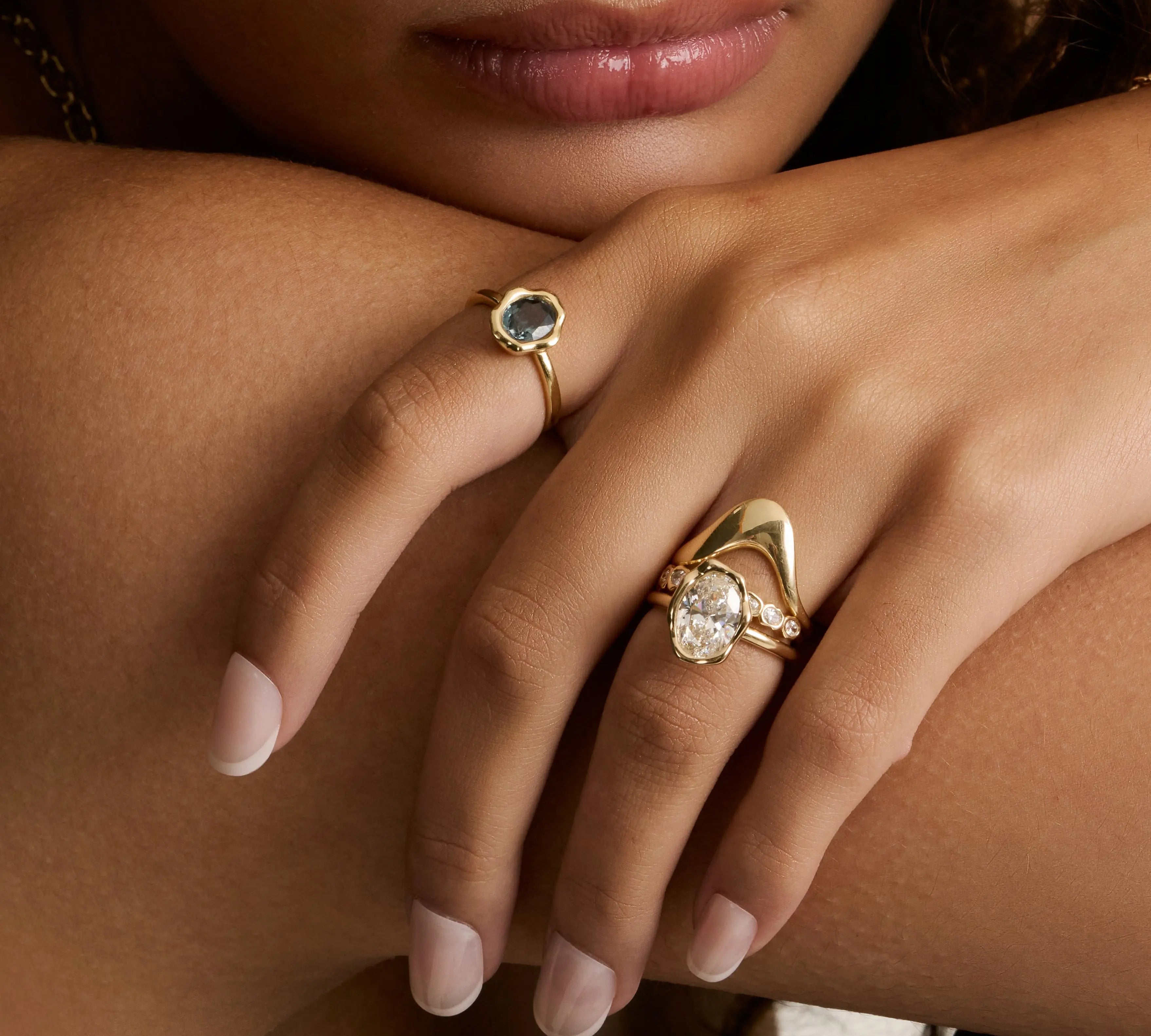 A close-up lifestyle shot of a model's hand resting on her arm, showcasing two gold rings. One ring features a round dark green stone, and the other features an oval diamond in a gold setting with a curved / asymmetrical accent.