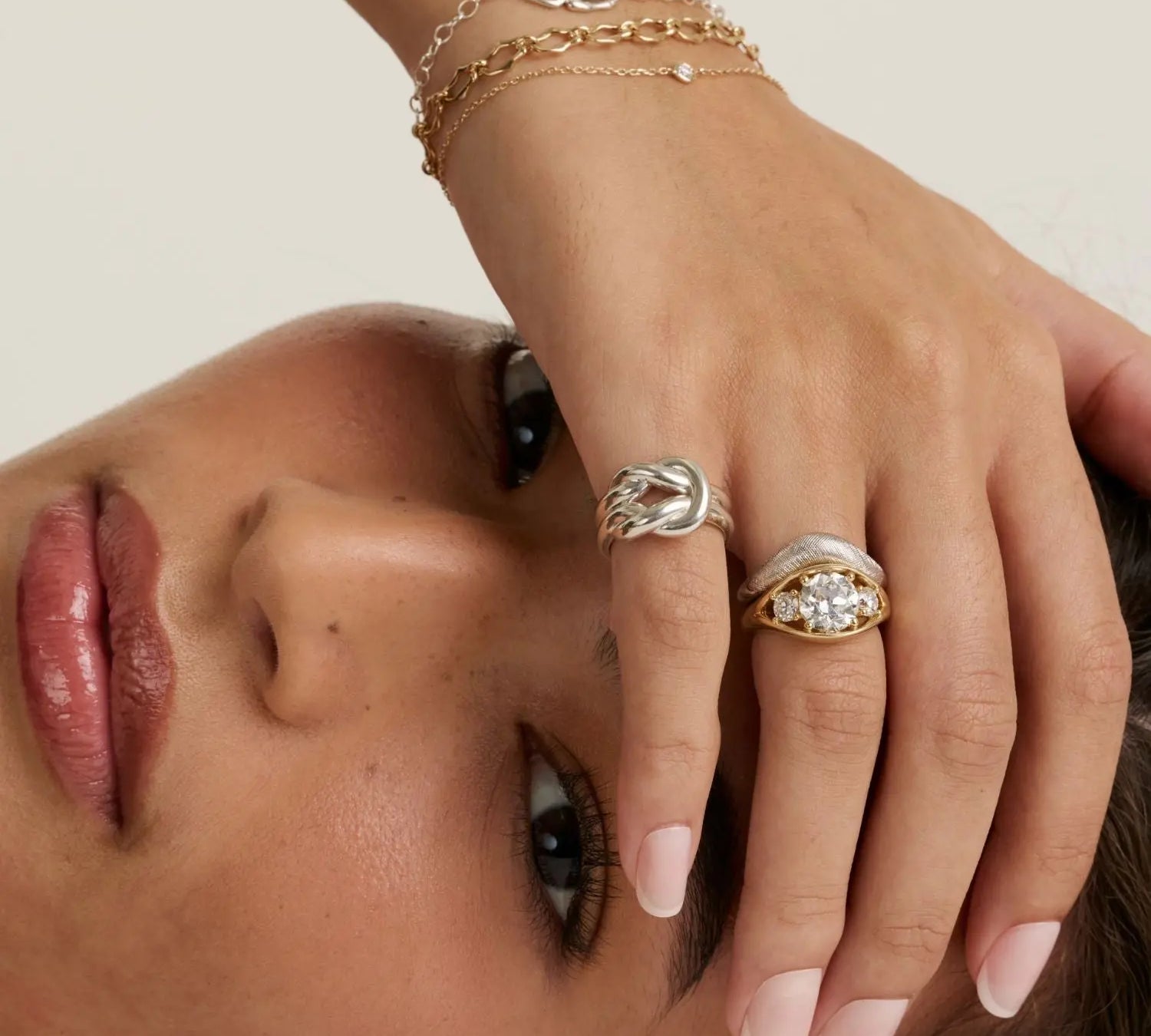 A close-up lifestyle shot of a model's hand wearing multiple rings, including a silver ring with a twisted knot design and a gold ring with a clear gemstone cluster, against a warm beige background.