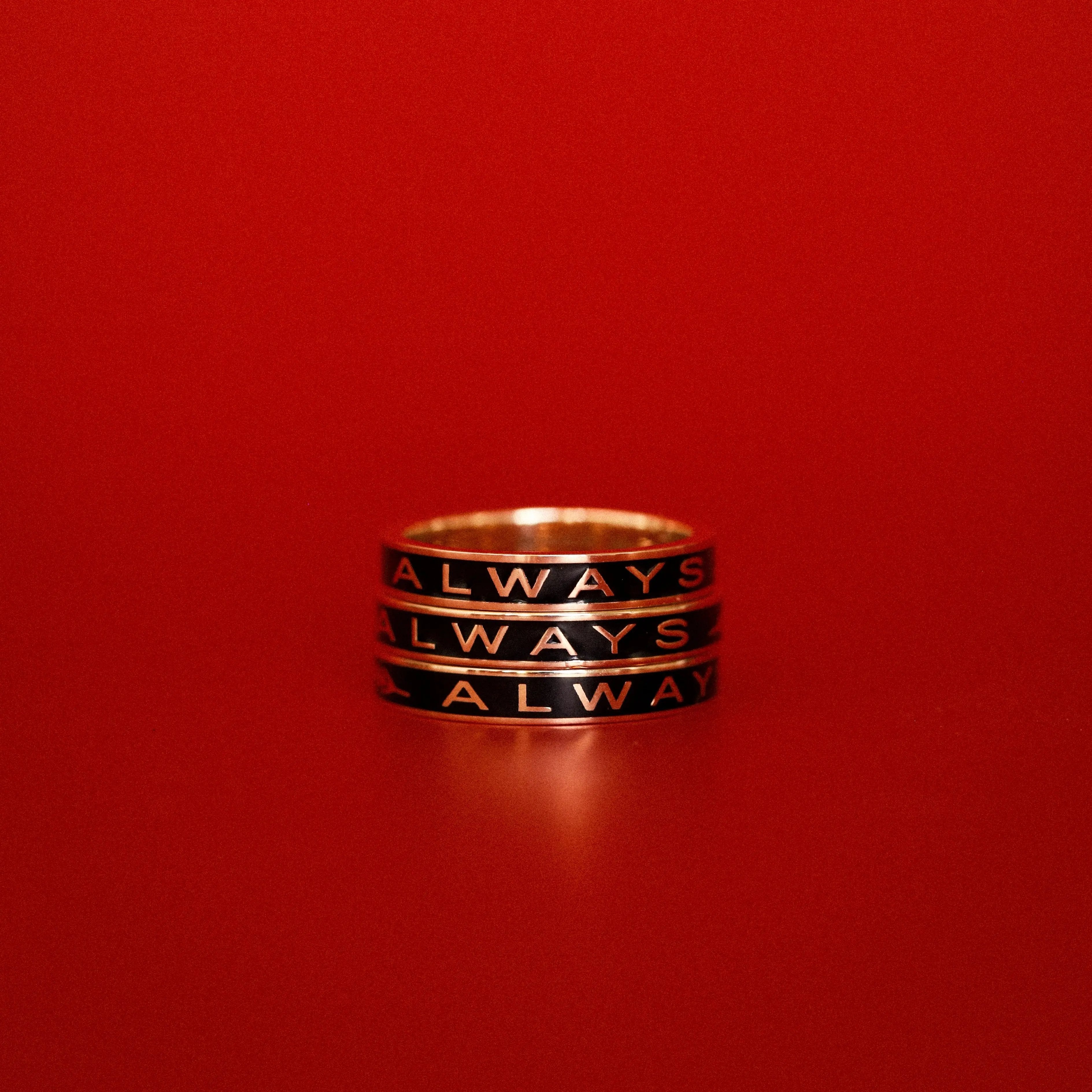 Three black enamel Always Gold Memory Rings stacked on top of each other against a red backdrop