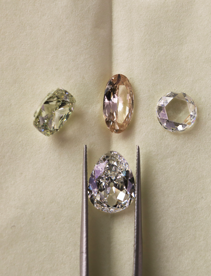 Four loose gemstones arranged on a beige textured surface, with a pair of metal tweezers holding a large pear-shaped clear gemstone below. The stones include a pale yellow oval, a peach oval, a round clear stone, and the large pear-shaped clear stone held in tweezers.