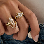 A close-up of a hand wearing multiple gold rings, including a ring with a large oval clear gemstone and another with a round clear gemstone, paired with blue denim jeans.