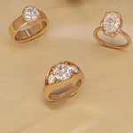 Three gold rings with clear gemstones in different shapes, arranged on a beige textured background.