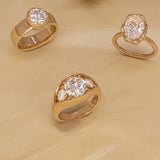 Three gold rings with clear gemstones in different shapes, arranged on a beige textured background.