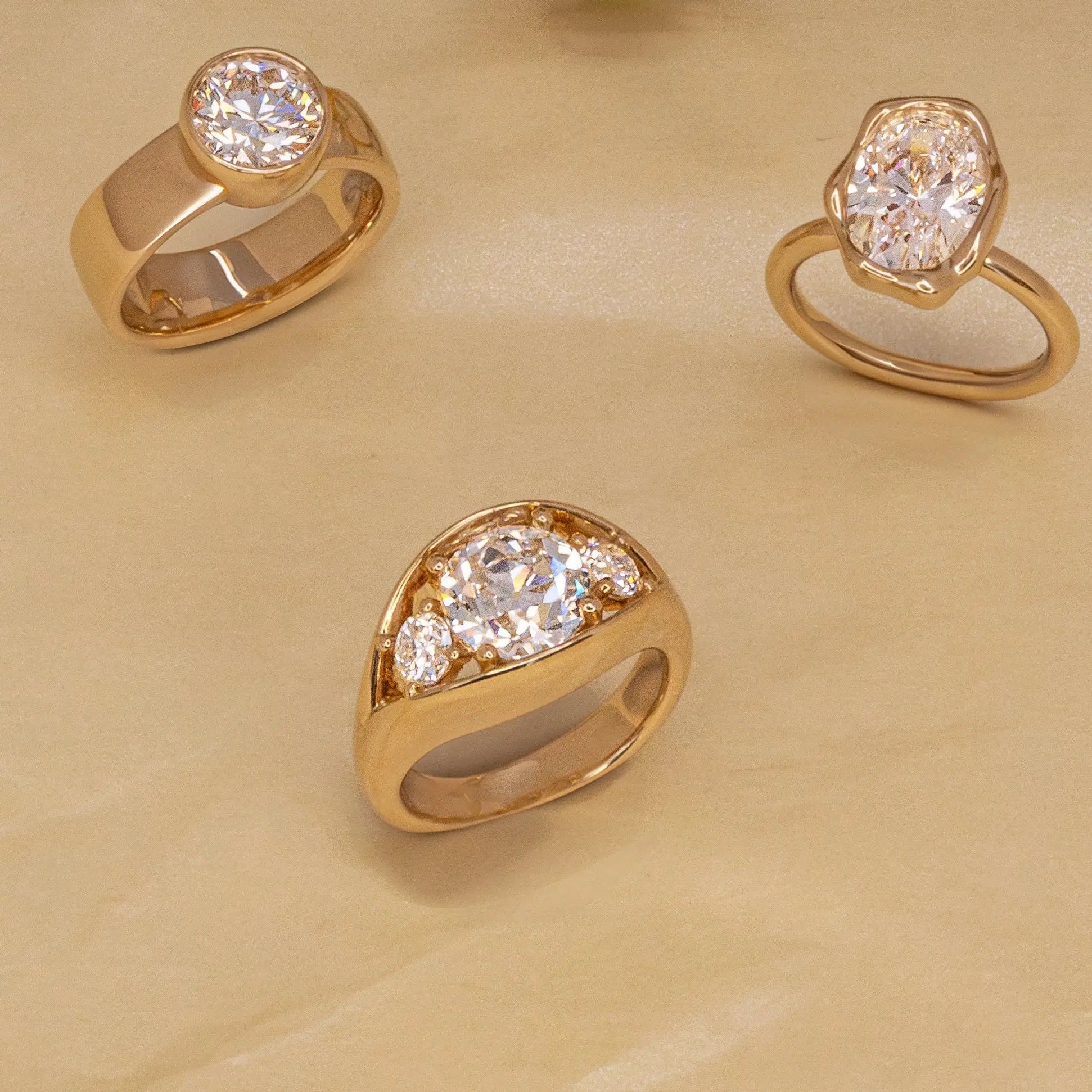Three gold rings with clear gemstones in different shapes, arranged on a beige textured background.