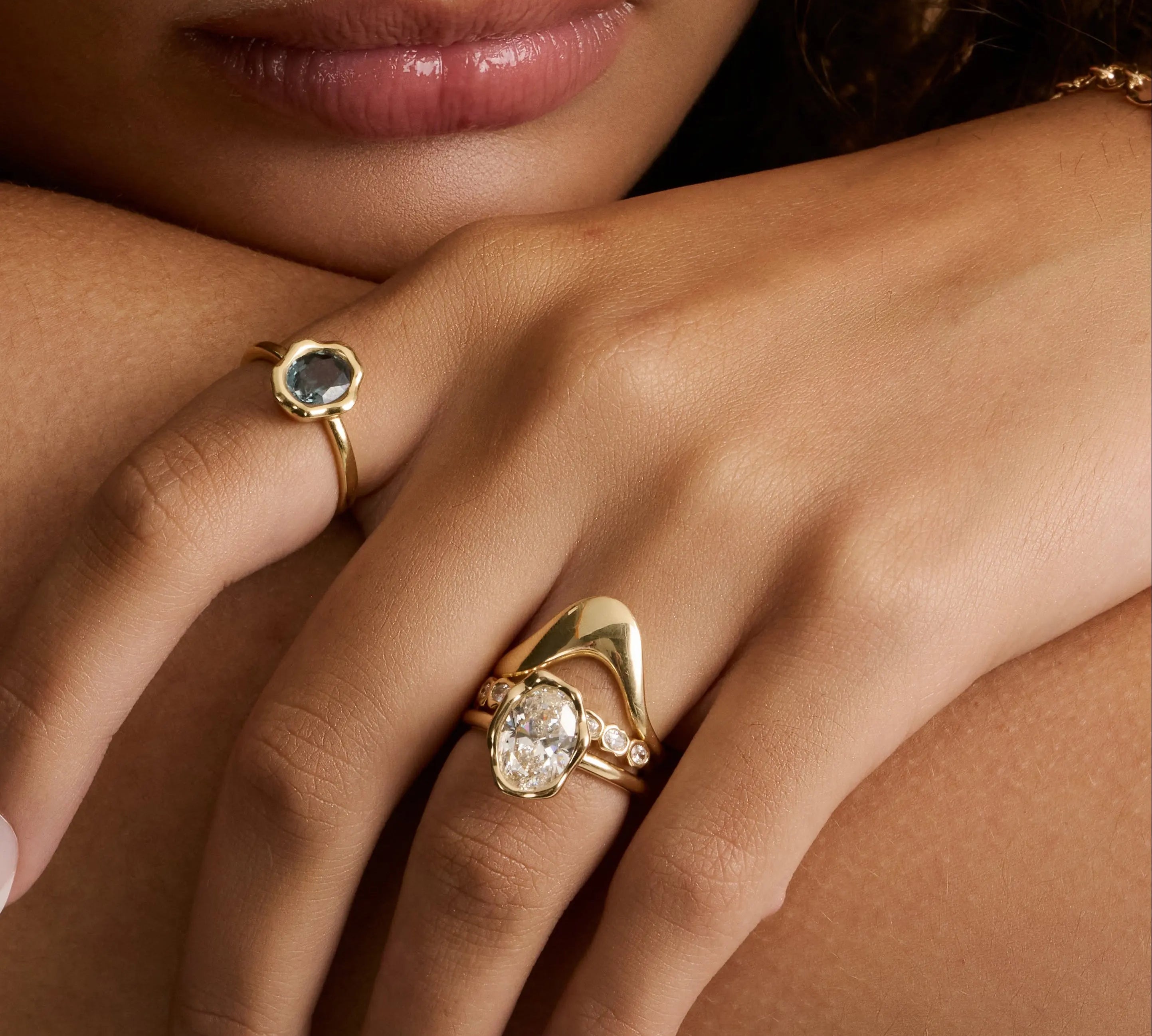A close-up lifestyle shot of a model's hand resting on her shoulder, showcasing two gold rings. The ring on her middle finger features a large, oval-cut clear gemstone, while the ring on her index finger has a smaller round stone with a dark blue-gray center and a textured gold setting.