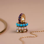 A gold charm with stacked rows of colorful rhinestones, including purple, turquoise blue, white, and clear stones, displayed on a beige background with a gold chain and partial bangle nearby.