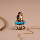 A gold charm with stacked rows of colorful rhinestones, including purple, turquoise blue, white, and clear stones, displayed on a beige background with a gold chain and partial bangle nearby.