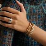 A close-up of a person's hand and wrist wearing gold jewelry, including a ring with green stones and stacked gold bracelets, against a patterned blue and brown top.