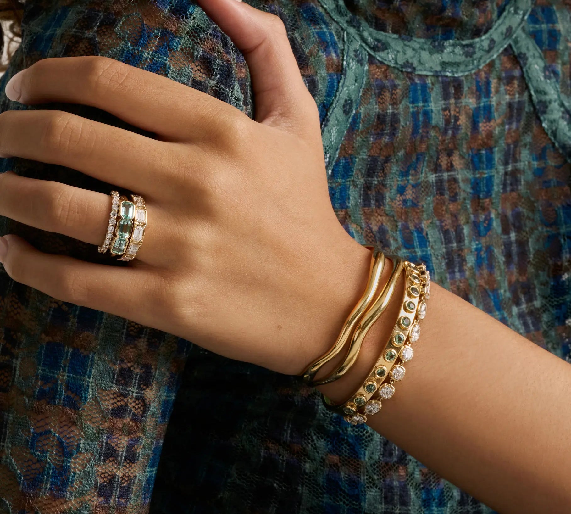 A close-up of a person's hand and wrist wearing gold jewelry, including a ring with green stones and stacked gold bracelets, against a patterned blue and brown top.