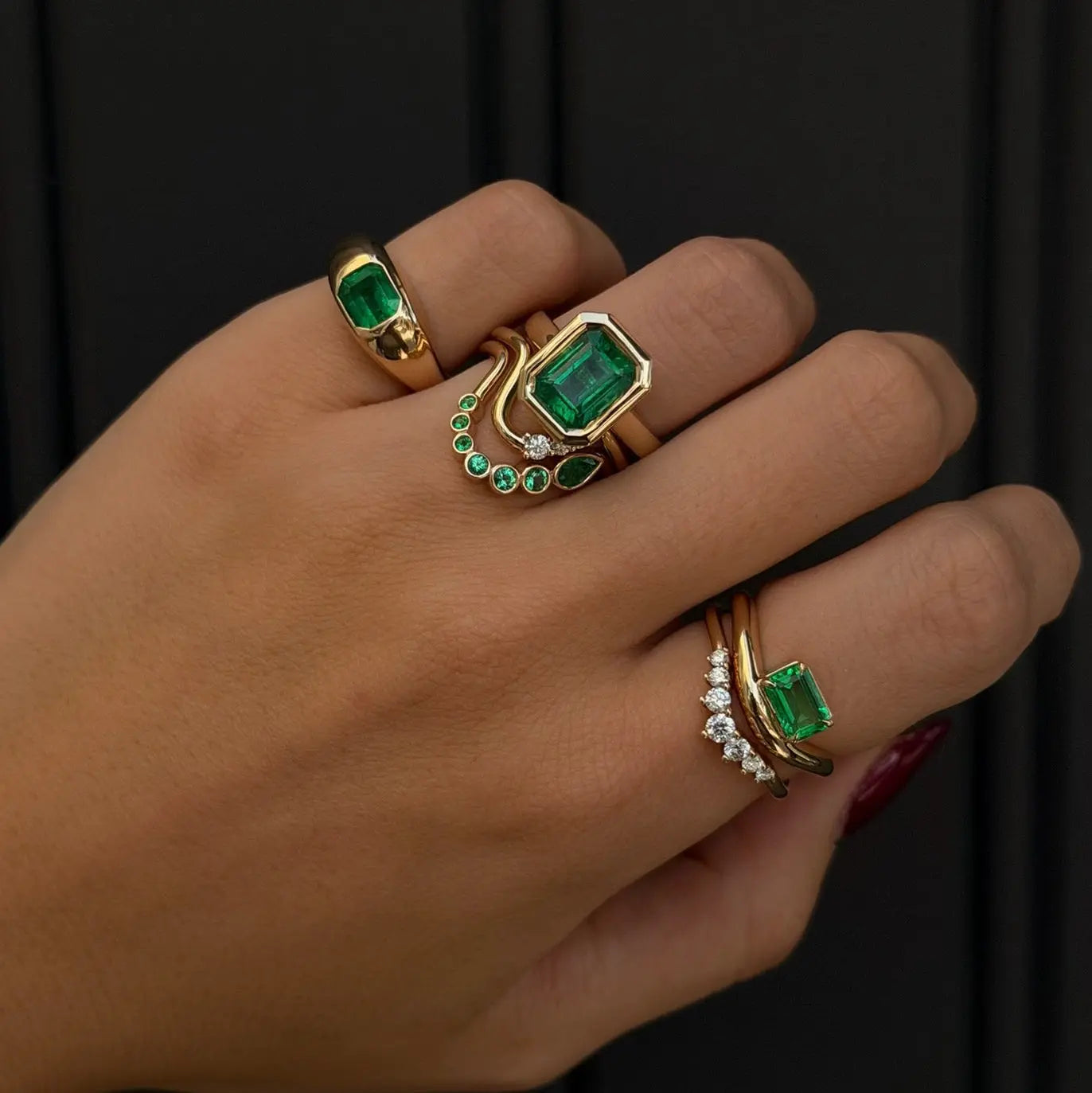 Hand wearing multiple gold rings with emerald green gemstones on a dark background