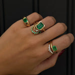 Hand wearing multiple gold rings with emerald green gemstones on a dark background