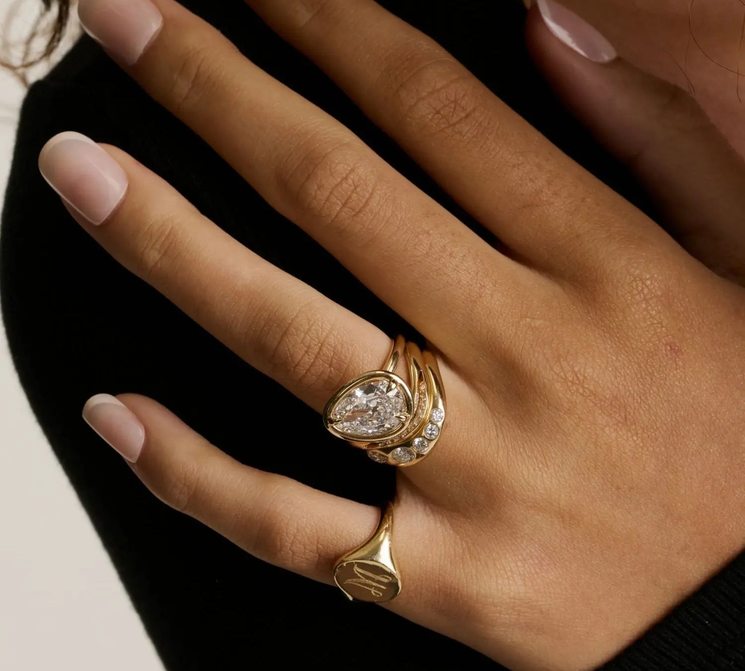 A close up of woman's hand wearing multiple yellow gold rings - an initial signet on the pinky and a tilted pear diamond engagement ring stacked with other wavy bands on wedding finger.