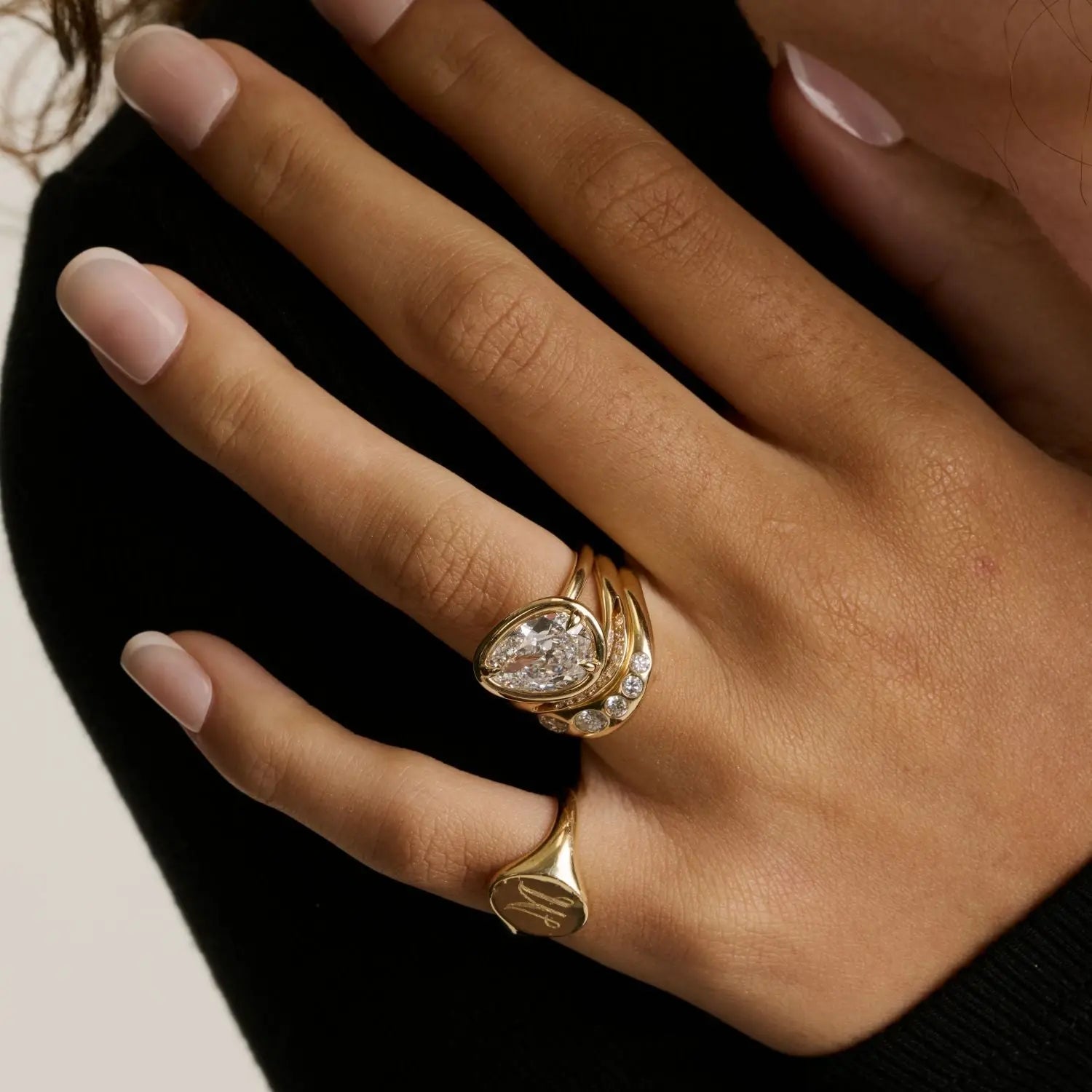 A close up of woman's hand wearing multiple yellow gold rings - an initial signet on the pinky and a tilted pear diamond engagement ring stacked with other wavy bands on wedding finger.