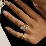 A close up of woman's hand wearing multiple yellow gold rings - an initial signet on the pinky and a tilted pear diamond engagement ring stacked with other wavy bands on wedding finger.