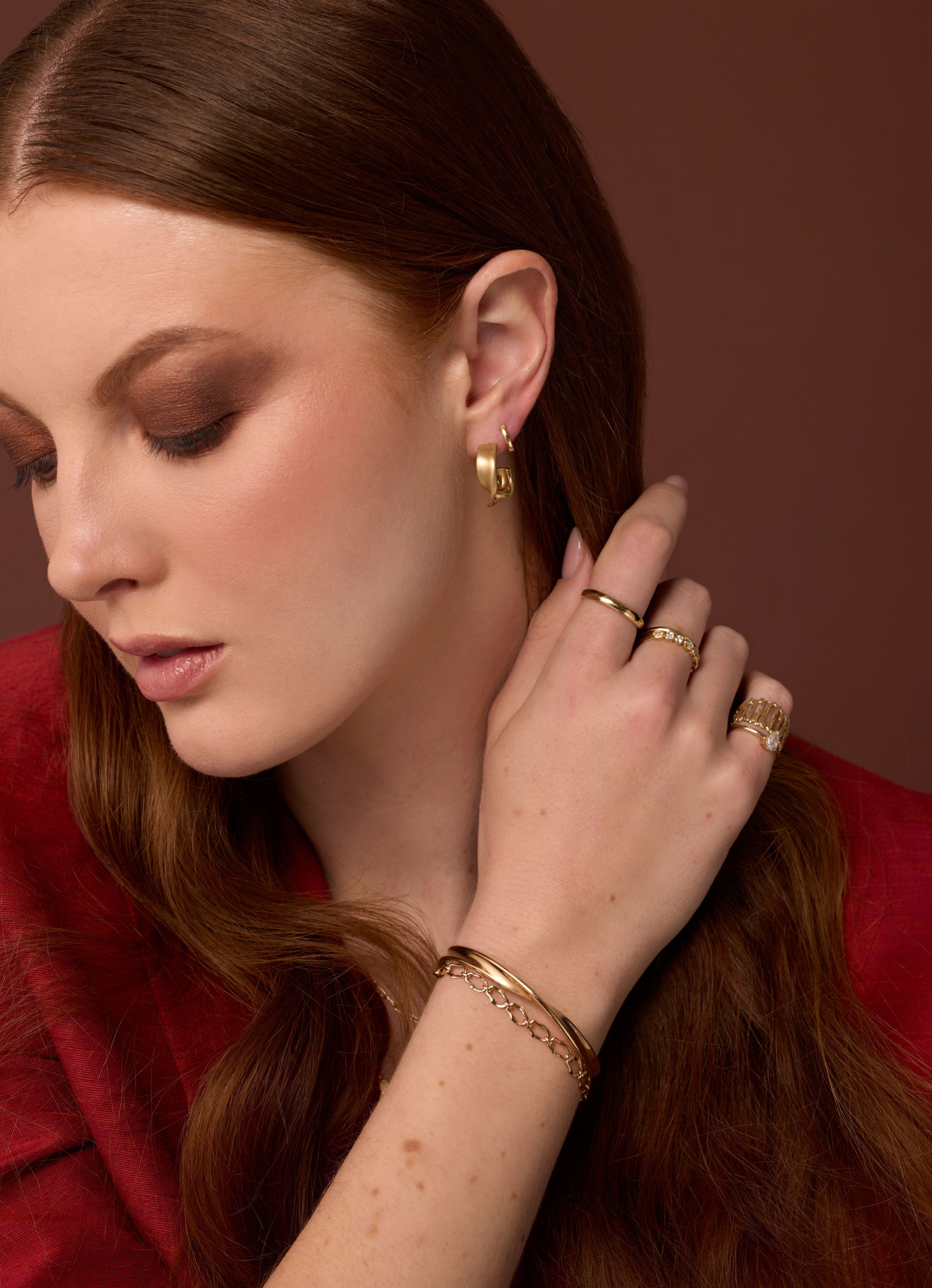 woman looking down wearing a red blazer against a dark maroon background wearing gold bracelets, rings and earrings
