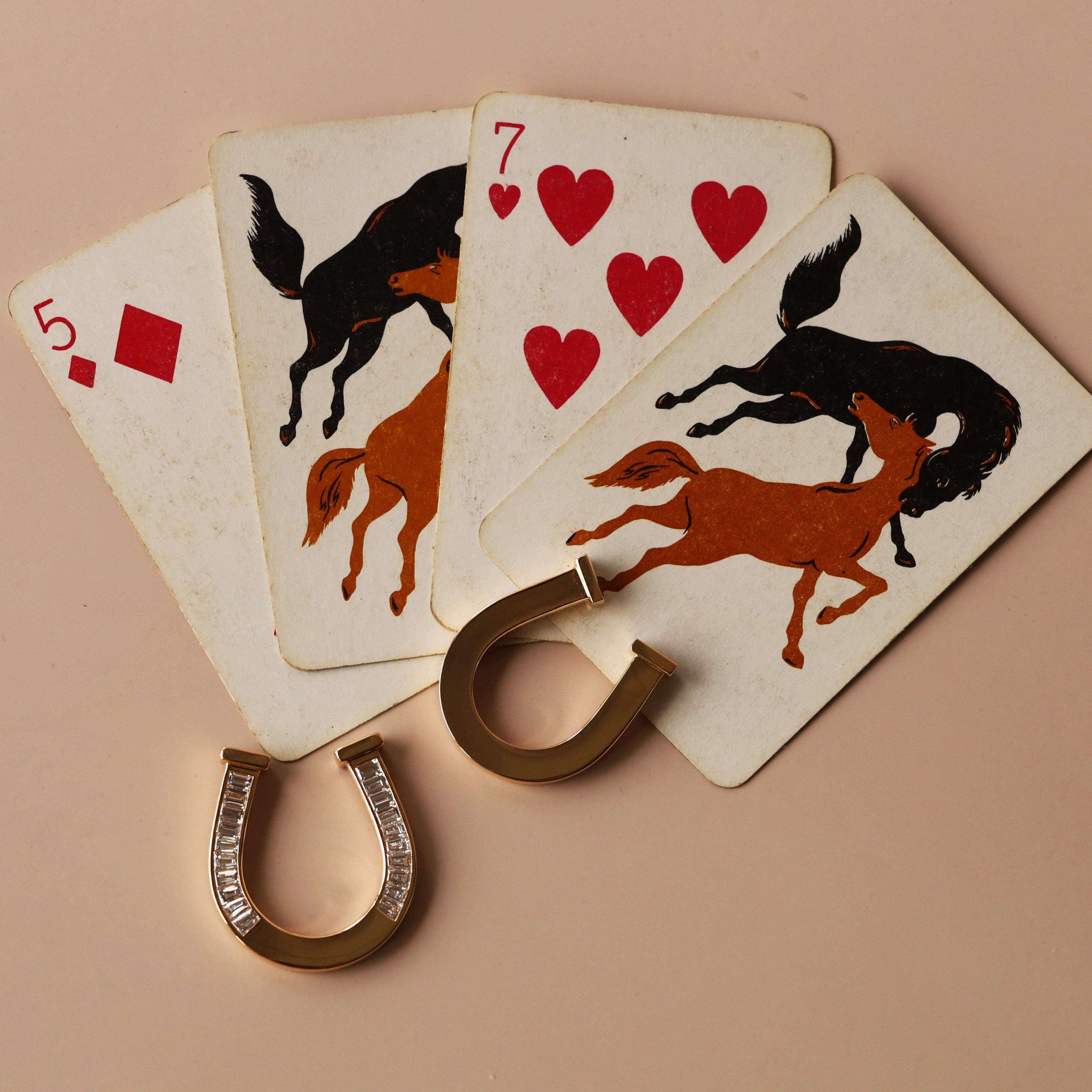 A set of four playing cards fanned out on a beige background, each featuring a horse and rider illustration in black and rust orange with red hearts. Two gold horseshoe-shaped accessories with clear rhinestones are placed in front of the cards.