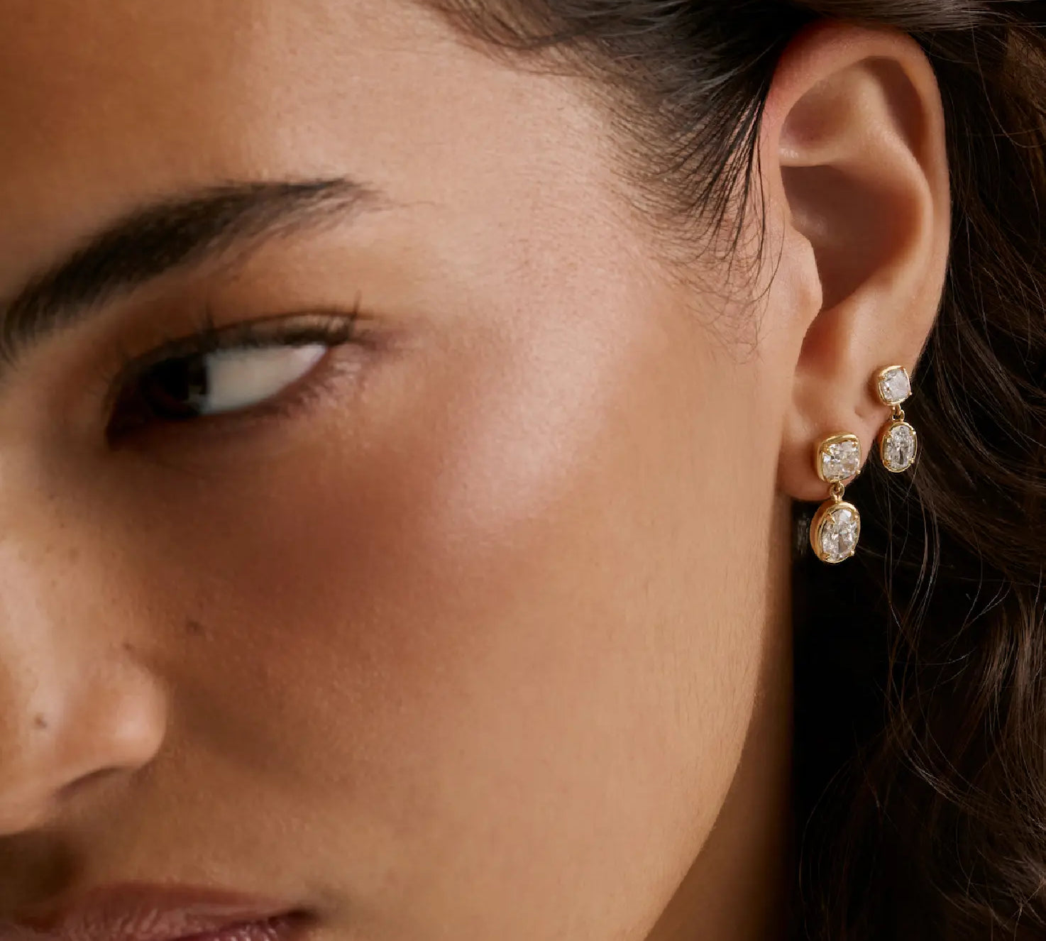 Close-up of a woman's ear wearing a pair of gold drop earrings with clear gemstones, shown against her skin and dark hair.