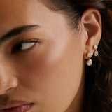 Close-up of a woman's ear wearing a pair of gold drop earrings with clear gemstones, shown against her skin and dark hair.