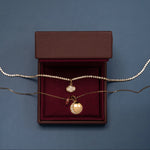 Margaret Tennis Necklace, Lisse East-West Oval Charm, moon phase charm and Birthstone Rondelle displayed on a burgundy jewelry box against a slate blue background