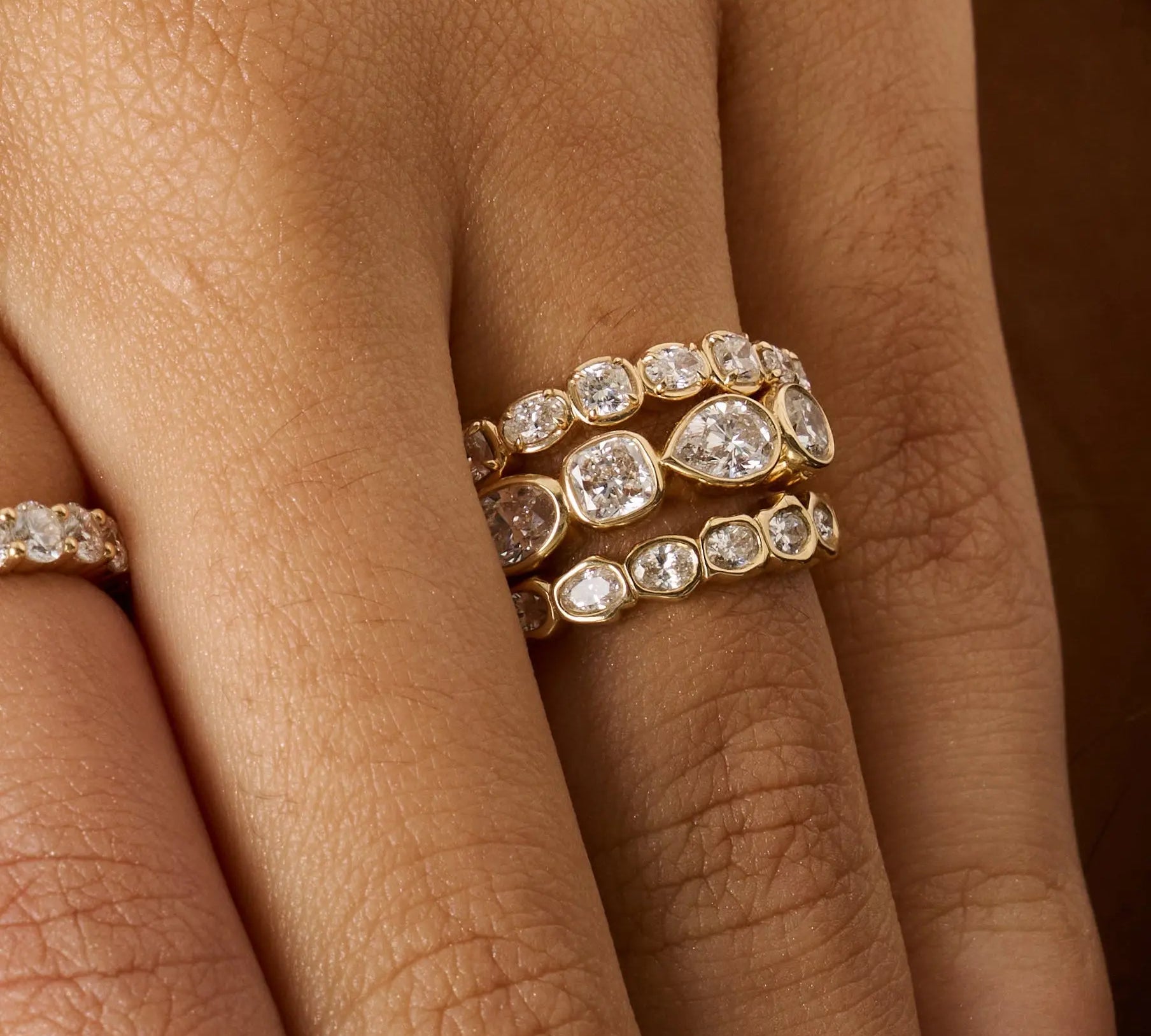 Close-up of a hand wearing three gold eternity rings set with clear round and pear-cut diamonds, with a fourth ring partially visible on the adjacent finger.