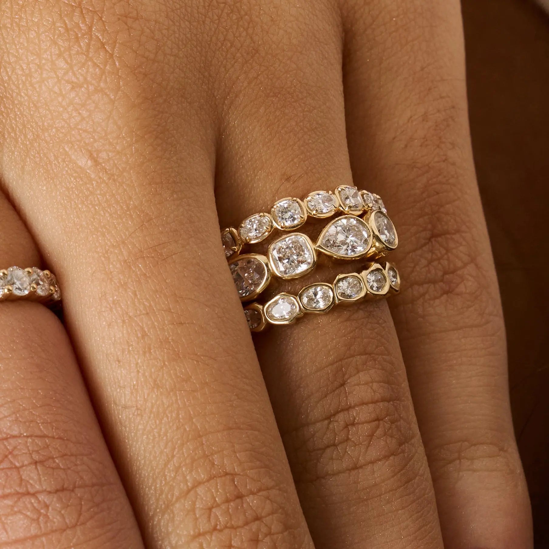 Close-up of a hand wearing three gold eternity rings set with clear round and pear-cut diamonds, with a fourth ring partially visible on the adjacent finger.