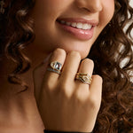 Close-up of a woman's hand wearing multiple rings, including a prominent gold ring with a square-cut clear gemstone and two stacked gold bands with geometric detailing, against a soft beige background.