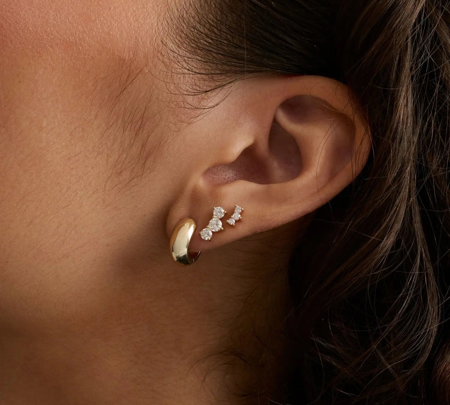 A close-up of a person's ear wearing three small gold earrings, including a huggie hoop and two delicate stud earrings with diamonds, against dark hair and warm skin tones.