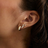 A close-up of a person's ear wearing three small gold earrings, including a huggie hoop and two delicate stud earrings with diamonds, against dark hair and warm skin tones.