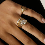 A close-up of a hand wearing multiple gold rings, including a signet-style ring and a stacked ring with a pear-cut clear gemstone and smaller round stones, against a dark background.