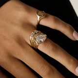 A close-up of a hand wearing multiple gold rings, including a signet-style ring and a stacked ring with a pear-cut clear gemstone and smaller round stones, against a dark background.