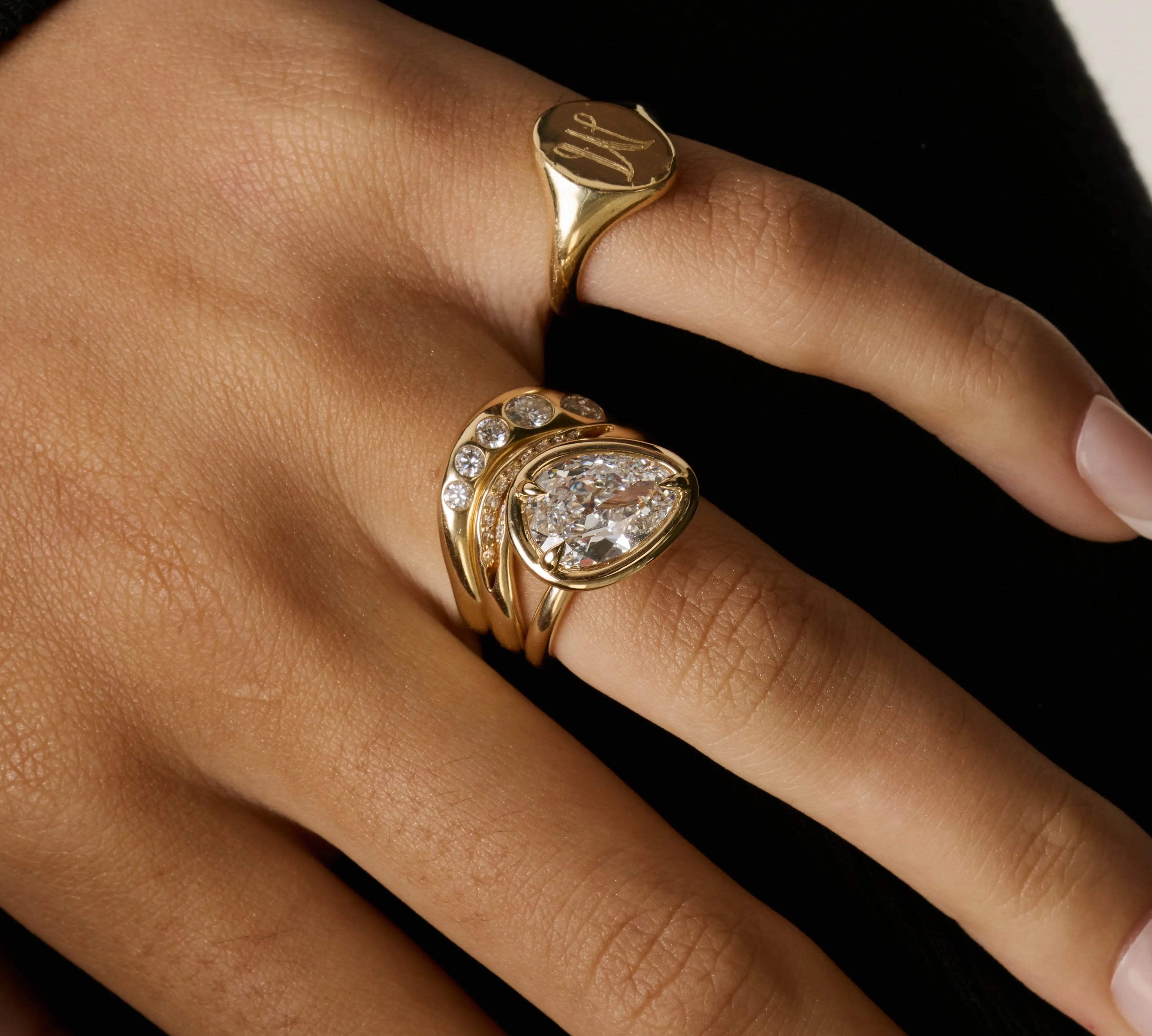 A close-up of a hand wearing multiple gold rings, including a signet-style ring and a stacked ring with a pear-cut clear gemstone and smaller round stones, against a dark background.
