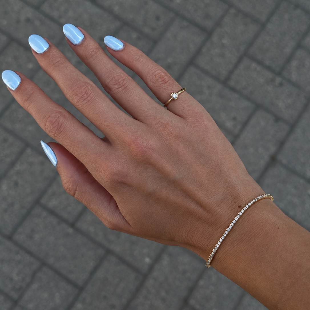 A close-up of a person's hand with light blue manicured nails, wearing a delicate gold tennis bracelet and a slim gold ring with a small clear gemstone, set against a gray paved background.
