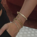 A close-up of a wrist wearing a stack of four gold bracelets: the Little White Diamond Cuff, the 8mm Sculpted Link Gold Chain bracelet, the Little Black Cuff, and the Mini Lisse Tennis Bracelet closest to the hand. The hand rests against black trousers as the wrist turns gently and the diamonds catch the light, with a deep red velvet chair in the background