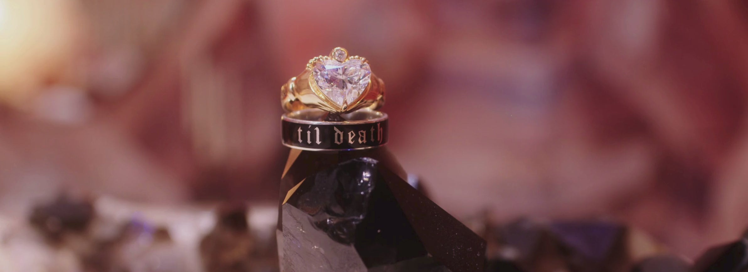 a black enamel til death ring placed on top of a black crystal, with a heart-shaped yellow gold engagement ring on top, subtle light movement against a blurry pink/red background
