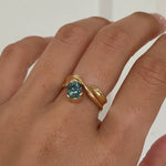 Hand wearing a gold ring with a blue gemstone on a plain background