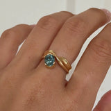Hand wearing a gold ring with a blue gemstone on a plain background